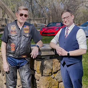 Chase L. Miller and Monte L. Miller posing in front of their sports cars, Monte wearing a leather biker's jacket and Chase wearing a vest and tie