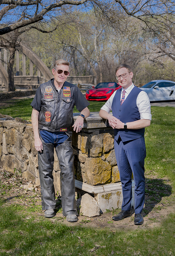 Chase L. Miller and Monte L. Miller posing in front of their sports cars, Monte wearing a leather biker's jacket and Chase wearing a vest and tie