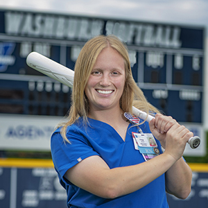 Paige Robbins posing on the softball field wearing nursing scrubs